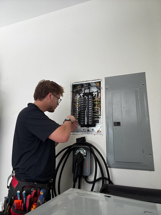 Amped Up electrician working at an open load center beside a newly installed EV charger, sizing the service for added load